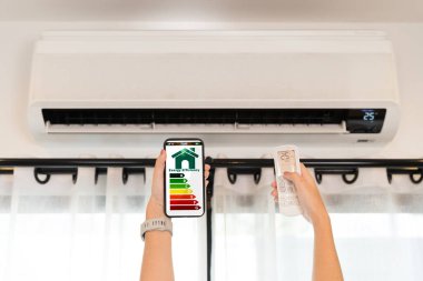 Akıllı telefonu tutan eller enerji verimliliğini ve evde uzaktan kumandalı klimayı gösteriyor elektrik tasarrufu akıllı yaşam teknolojisi sürdürülebilir yaşam tarzını konforlu kılıyor.