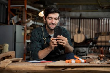 Önlüklü marangoz marangoz marangozluk, ahşap atölyesindeki bir tezgahta akıllı telefon kullanır. Modern zanaat işlerini, müşteri mesajlarını, online siparişleri ve proje planlamasını gösterir..