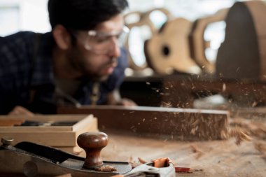 Bir marangoz tozu savururken ahşap talaşları uçuşan bir tezgahın üzerindeki el uçağının yakın çekimi. Atölye araçları ve arka planda bulanık zanaatkar, marangozluk ve zanaat kavramı.