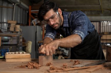 Güvenlik gözlüğü takan odaklanmış bir marangozun yakın plan resmi. Tezgahta el uçağını ahşap üzerinde kullanıyor. Bir atölyede ahşap talaşı ve aletler, zanaat ve marangozluk kavramı.