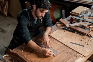 Odaklanmış luthier, aletler ve parçalarla çevrili ahşap tezgah üzerinde gitar eğrilerinin izini sürmek için bir kalem ve temiz bir şablon kullanır. Hassasiyet tasarımı, ahşap işçiliği ve onarımını gösterir..