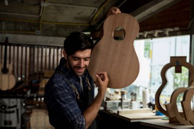 Gülümseyen Luthier bitmemiş bir akustik gitarı bir marangozhanede boş tutuyor. El yapımı müzik aletleri, ahşap işçiliği, zanaatkar üretimi ve küçük işletme atölyesi kavramı.