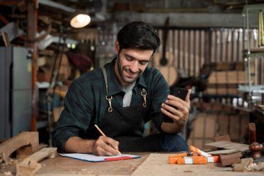 Önlüklü gülümseyen marangoz akıllı telefonu kontrol ederken çalışma masasındaki panoya notlar yazıyor. Atölyede küçük işletme, zanaat, planlama ve modern teknoloji kavramı.