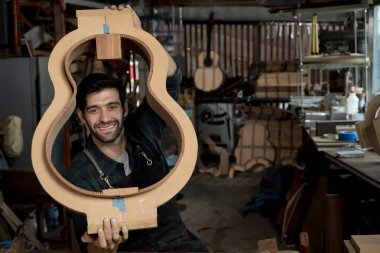 Happy gitarist Luthier, yoğun bir atölyede yüzünü çerçeveleyen tahta gitar kalıbını gösteriyor. El yapımı enstrüman, alet ve şablon ile sanatsal portresi.