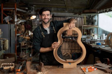 Gülümseyen gitarcı bir marangoz atölyesinde tezgahın üzerinde gitar gövdesi kalıbı tutuyor. El yapımı enstrümanlar için araçlar, kıskaçlar ve şablonlar ile zanaatkar portresi.
