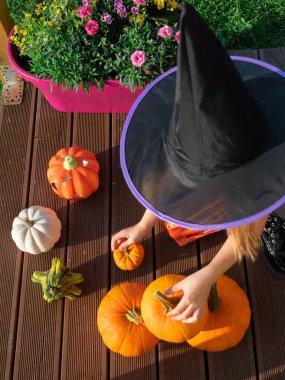 Cadı kostümlü kız tahta terasta balkabaklarıyla oynuyor.