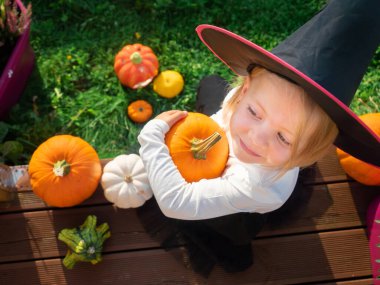 Cadı kostümlü kız tahta terasta balkabaklarıyla oturuyor.