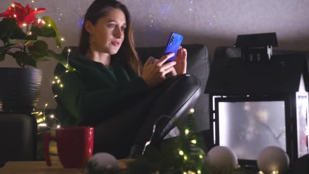 Jovem mulher sentada no sofá no Natal decorado sala de estar usando telefone celular. — Vídeo de Stock