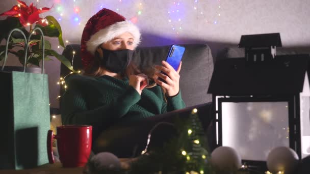 Молодая женщина в шляпе Санты и черной маске с помощью мобильного телефона сидит дома — стоковое видео