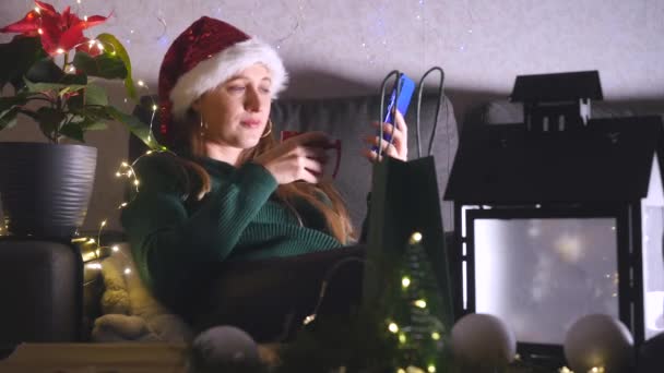 Mladá žena v santa klobouk sedí na pohovce pití čaje a pomocí mobilního telefonu. — Stock video