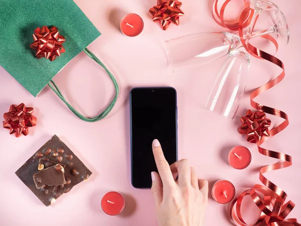 Bir kadın akıllı telefon ekranına dokunuyor. Sevgililer Günü geçmişi. Hediye, mumlar, pembe arka planda gözlük.