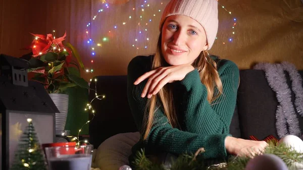 Jovem mulher de chapéu de malha sentada no quarto decorado de Natal sorrindo para a câmera — Fotografia de Stock