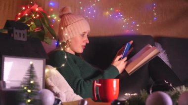 Noel süslemeli oturma odasında koltukta oturan genç bir kadın cep telefonu kullanıyor.. 