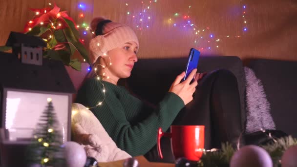 Junge Frau mit Kopfhörer und Hut sitzt auf dem Sofa im weihnachtlich dekorierten Wohnzimmer und benutzt Handy. — Stockvideo