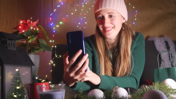 Şapkalı genç kadın akıllı bir telefon tutuyor ve Noel arifesinde video görüşmesi yapıyor. — Stok video