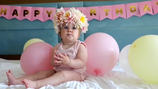 Αξιολάτρευτο κοριτσάκι με στεφάνι από λουλούδια να κάθεται στο κρεβάτι με μπαλόνια στα πρώτα της γενέθλια. — Αρχείο Βίντεο