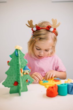 Geyik boynuzu takan genç bir kız, plastik bir Noel ağacı için renkli kil süslemeleri yapmaktan hoşlanıyor.