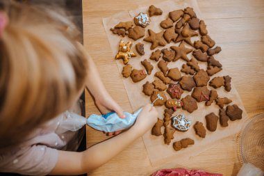 Bir çocuk mutfak masasında çeşitli zencefilli kurabiyeleri kremalarken görülüyor. Renkli dekorasyonlar uygulanıyor.