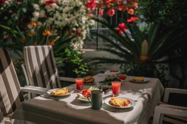 Bahçedeki bir masada iki tabak yiyecek ve içecek duruyor. Sandalyeler yakınlarda ve etrafında çiçekler var..
