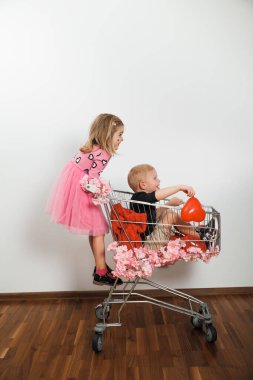 İki çocuk, sade bir odada çiçeklerle süslenmiş bir alışveriş arabasında eğleniyorlar..