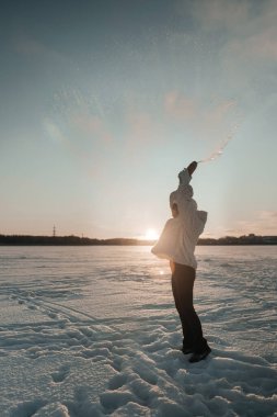 Bir insan donmuş bir gölde duruyor ve gün batımında havaya kar atıyor..