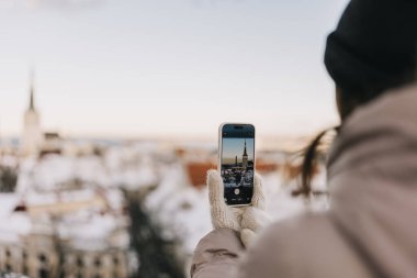 Bir insan elinde telefonla karda durup güneşli bir günde arka planda bir kuleyle Tallinn Eski Şehir manzarasını yakalıyor..