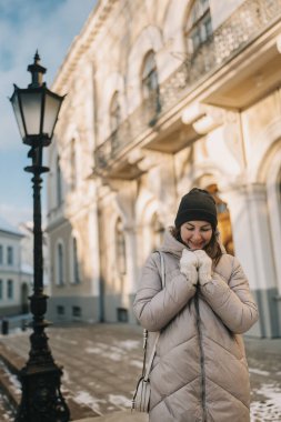 Bir kadın, kışın tarihi bir binanın önünde elini sıcak ve neşe için tutarken gülümsüyor..