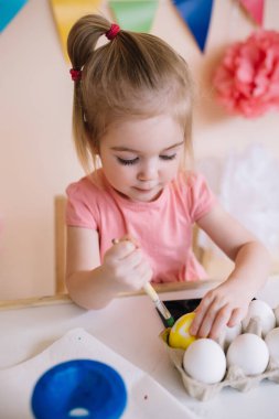 Sevimli mutlu 3 yaşında pembe elbiseli bir kız Paskalya yumurtalarını parlak renkli bir odada boyuyor.