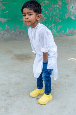 Gülümseyen bir çocuk modern bir elbise olarak beyaz Punjab ve sarı ayakkabılı mavi kot pantolon giyen bir fotoğraf için poz veriyor. İki yaşında bir çocuk, meraklı bir suratla uzaklara bakıyor..