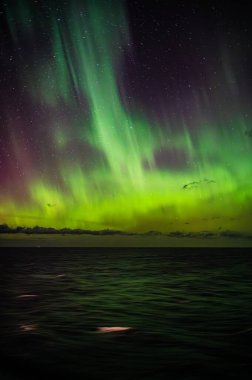 Kuzey Işıkları 'nın büyüleyici bir görüntüsü, gece gökyüzünü dingin bir okyanusun üzerinde aydınlatır. Suyun üzerinde canlı yeşil ve mor bir parıltı bırakır.
