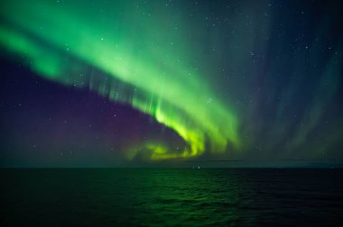 Kuzey Işıkları 'nın büyüleyici bir görüntüsü engin bir okyanus üzerinde gece gökyüzünü aydınlatarak büyüleyici, yeşil ve mor bir aurora yaratıyor.