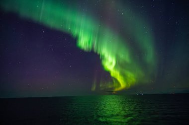 Kuzey Işıkları 'nın büyüleyici bir görüntüsü gece gökyüzünü okyanus üzerinde aydınlatır. Karanlık zemine karşı canlı, ışıldayan bir yay yaratır.