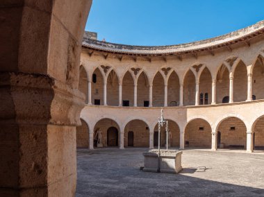 Castell de Bellver. Palma de Mallorca, tarihi dairesel avlu merkezinde, üzerinde açık mavi gökyüzü olan kemerli sütunlarla çevrili taş bir çeşme bulunur, Majorca.