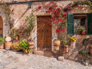 Valldemossa, Mallorca, canlı kırmızı çiçekler ve yeşil panjurlarla süslenmiş büyüleyici bir taş bina, saksı bitkileri ve tuhaf bir kaldırım taşı yolu olan Majorca 'nın çevresindeki hoş bir ahşap kapı içeriyor.