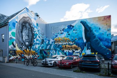 Bir binanın üzerinde canlı bir duvar resmi, bir adamın büyük bir portresi, uzanan bir el, ve önünde arabalar park etmiş mavi bir balina, Tromso, Norveç