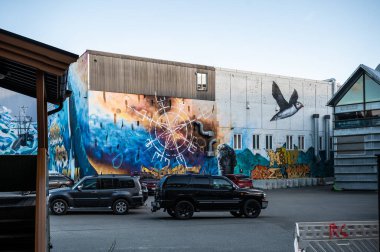 Binanın üzerindeki renkli bir duvar resminde, arka planda bir heykel ve deniz unsurları bulunan, göksel sembollerle çevrili, suyun üzerinde uçan bir kuş bulunur. Arabalar yakınlarda park halinde, Tromso, Norveç