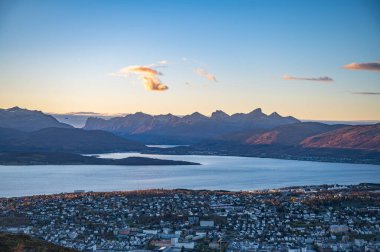Tromso, Norveç 'in panoramik manzarası, sakin bir göl ve görkemli dağlar arasına kurulmuş, şafağın ılık renklerinde, Storsteinen' in tepesinde.
