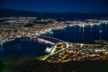 Tromso, Norveç 'in çarpıcı bir gece manzarası, ışıklarla aydınlatılmış, bir liman ve nehir boyunca uzanan bir köprü, Storsteinen' in tepesinde.
