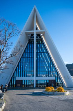Kuzey Kutbu Katedrali, Tromso, Norveç. Modern kilise, üç köşeli cam bir cephe ve büyük bir haç, mavi gökyüzü, birkaç kişi tarafından çevrelenmiş.