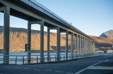 Tromsobrua Köprüsü, uzun köprü bir su kütlesi üzerinde uzanır, arka planda dağlar ve yolda görünen bir park işareti, Tromso, Norveç