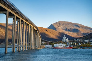 Tromsobrua Köprüsü, bir su kütlesi boyunca uzanır, kırmızı ve beyaz bir tekne, arka planda Arktik Katedrali, Tromso Norveç