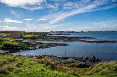 Kyststien, Haugesund, Norveç, sakin kıyı manzarası küçük kırmızı çatılı bir ev, sakin bir su kütlesi ve parçalı bulutlu bir gökyüzünün altında uzak bir sanayi bölgesi.
