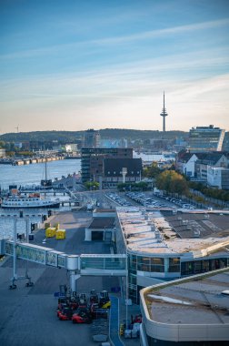 Kiel, Almanya 'nın hareketli liman sahnesi, feribot iskelesi, modern binalar, ve arka planda yüksek bir kule, berrak bir gökyüzünün altında, dikey çekim