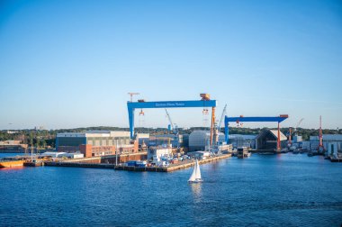 Alman Deniz Kuvvetleri, Kiel, Almanya, açık mavi gökyüzü altında hareketli bir limanda süzülüyor, arka planda büyük vinçler ve endüstriyel binalar var.