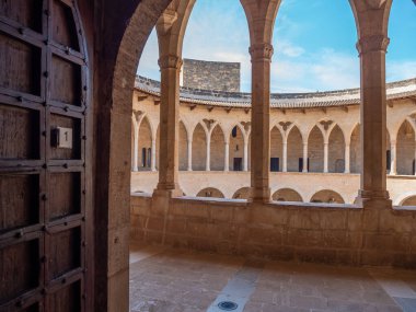 Castell de Bellver, Palma de Mallorca, ahşap bir kapıdan görünen manzara, arka planda mavi gökyüzünün altında kemerli sütunları ve taş bir binası olan açık bir avluyu ortaya çıkarıyor.