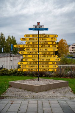Norveç 'in Narvik kentindeki bir yönlü sarı işaret direği Viyana, Rovaniemi ve diğer şehirler de dahil olmak üzere yakınlardaki bölgelere uzaklıkları ve yönleri gösteren çeşitli noktalar göstermektedir.