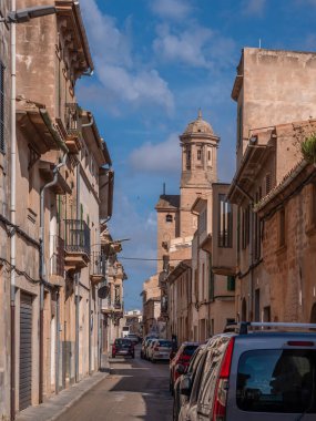 Llucmajor, Mallorca, dar sokaklar eski binalar ve park edilmiş arabalarla dolu. Arka planda tarihi bir kule görünüyor. Açık mavi gökyüzünün altında, Majorca.