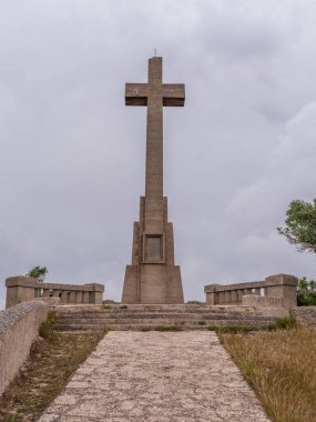 Puig des Mila, Sant Salvador, Mallroca, büyük taş haç heykeli bulutlu bir gökyüzünün altında, çimenler ve ağaçlarla çevrili bir taş yolun sonunda duruyor.