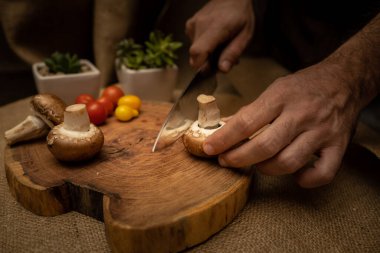 Ahşap bir tahtanın üzerinde Portobello mantarı doğrayan bir adamın eline yakın çekim. Çuval bezi kumaş arkaplanı.