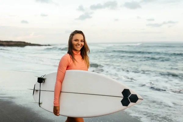 Mulher surfista atraente nova com placa branca no por do sol no oceano. Bali Indonésia. — Fotografia de Stock